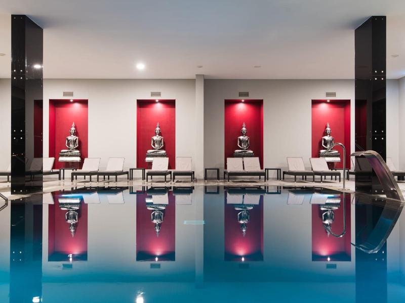 Moderner Innenpool mit ruhigem Wasser, vier Buddha-Statuen auf rotem Hintergrund und bequemen Liegen.