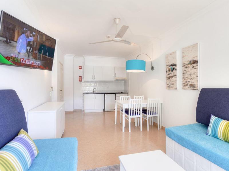 Helles Wohnzimmer mit zwei blauen Sofas, Esstisch, Küche und Wandfernseher in einer Ferienwohnung.