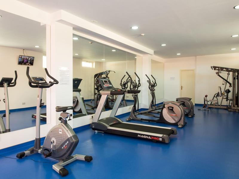 Heller Fitnessraum mit blauem Boden, mehreren Ergometern, Laufband und Krafttrainingsgeräten.