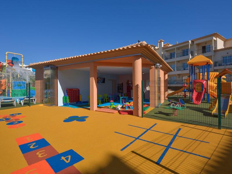 Bunter Kinderspielplatz mit Klettergerüst und Basketballkorb vor Gebäude bei klarem Himmel.