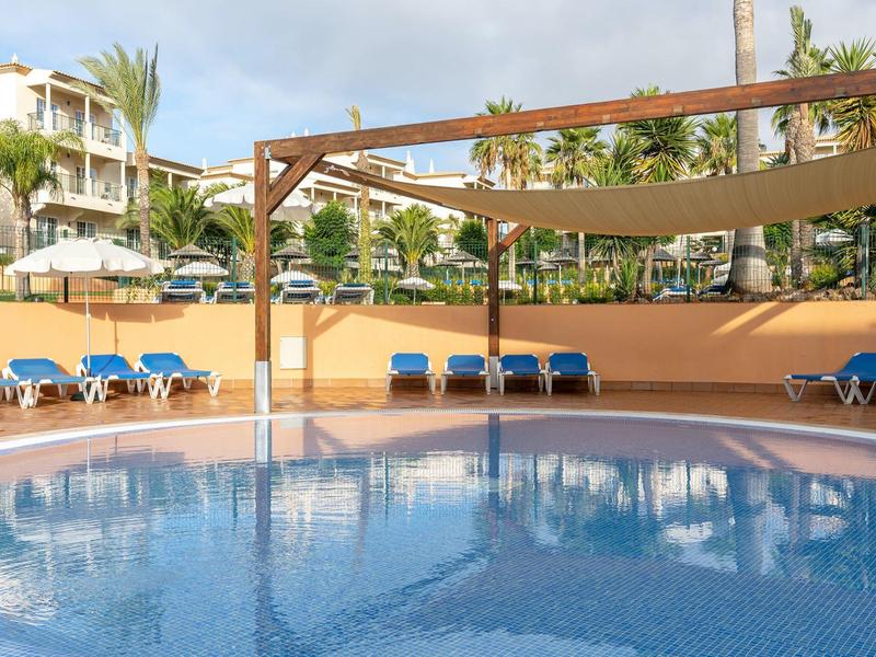 Hotelpool mit blauen Liegestühlen und Sonnensegeln unter bewölktem Himmel.