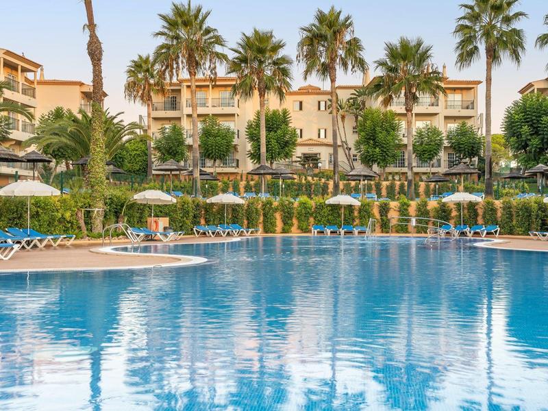 Großer Hotelpool mit Liegestühlen, Sonnenschirmen und Palmen vor einem mehrstöckigen Gebäude.