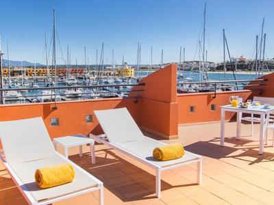 Terrasse mit orangen Wänden, zwei weißen Liegen, Tisch, Stühlen und Blick auf Hafen und Boote.