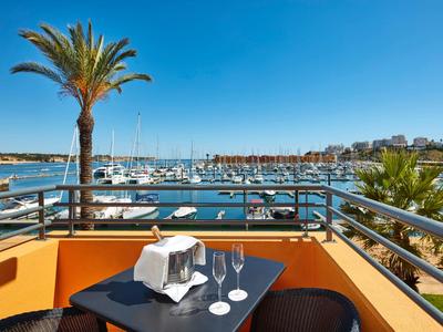 Balkon mit Tisch und Stühlen, Palmbaum und Blick auf Hafen mit Segelbooten und blauem Himmel.