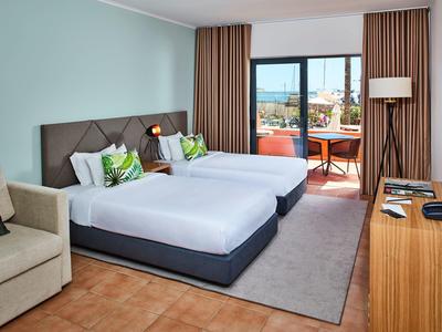 Helles Hotelzimmer mit zwei Betten, Balkon, braunen Vorhängen und Holzmöbeln.