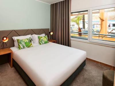 Modernes Schlafzimmer mit großem Doppelbett, grünen Kissen und Fenster mit Blick ins Freie.