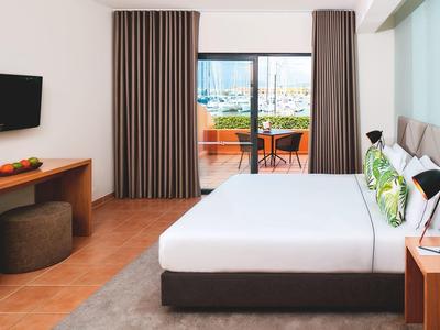 Hell eingerichtetes Schlafzimmer mit großem Bett, TV, Balkon und braunen Vorhängen.