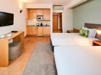 Hotelzimmer mit zwei Betten, Küchenzeile, Wand-TV und modernem, hellem Interieur.