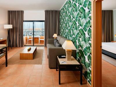 Modernes Hotelzimmer mit tropischer Wand, dunklem Sofa, Holzmöbeln und Balkon mit Meerblick.