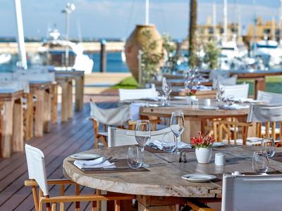 Freiluftrestaurant mit rundem Holztisch, weißen Stühlen und Blick auf den Hafen mit Booten.