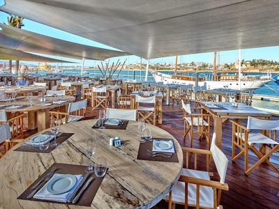 Offenes Restaurant mit Holztischen, weißen Stühlen und Blick auf Wasser und Stadt im Hintergrund.