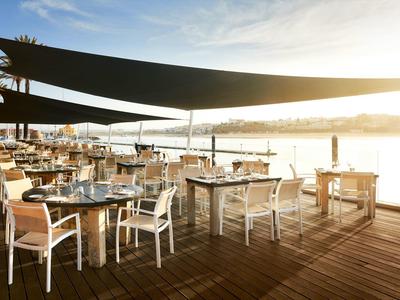 Sonnige Terrasse mit Holzboden, weißen Stühlen, Tischen und schwarzen Sonnensegeln am Wasser.