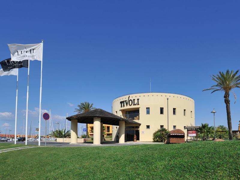 Rundes gelbes Gebäude "Tivoli" mit Palmen, grüner Wiese und blauen Himmel. Drei Fahnen wehen.