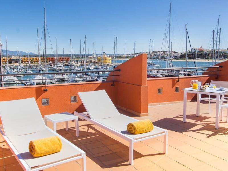 Terrasse mit orangen Wänden, zwei weißen Liegen, Tisch, Stühlen und Blick auf Hafen und Boote.