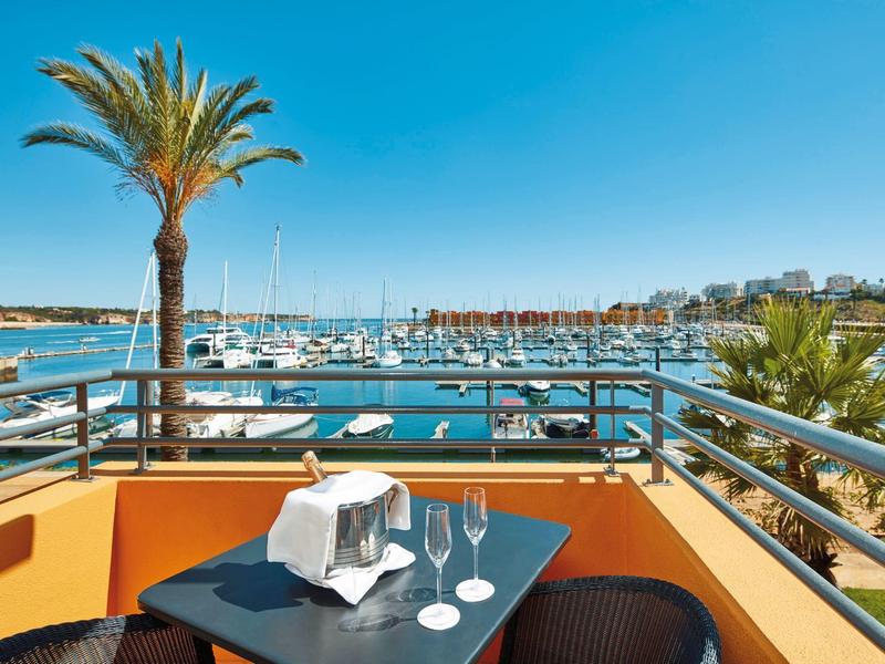 Balkon mit Tisch, Gläsern und Blick auf Hafen mit Booten, Palmen und Häusern im Hintergrund.