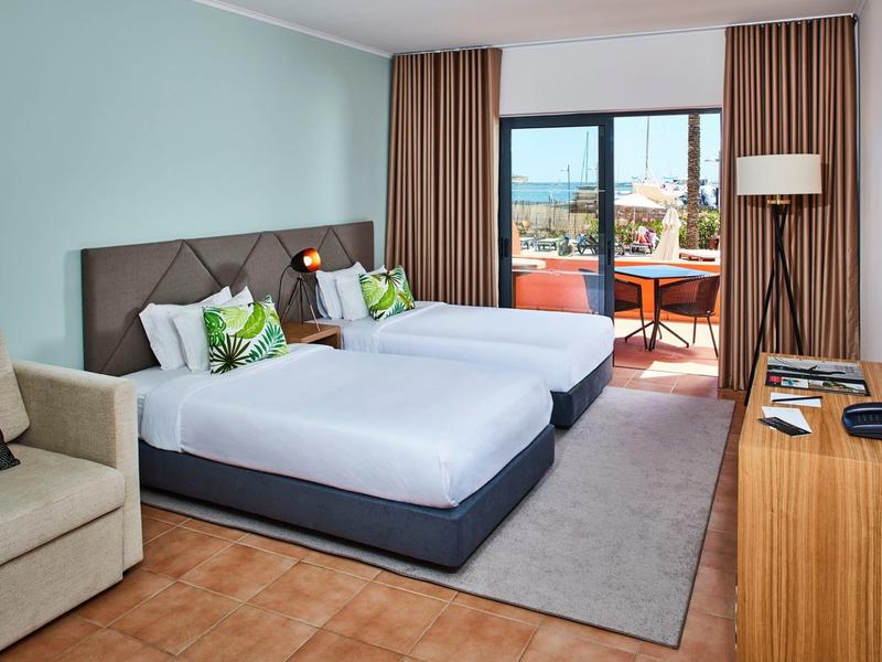 Helles Hotelzimmer mit zwei Betten, Balkon, braunen Vorhängen und Holzmöbeln.