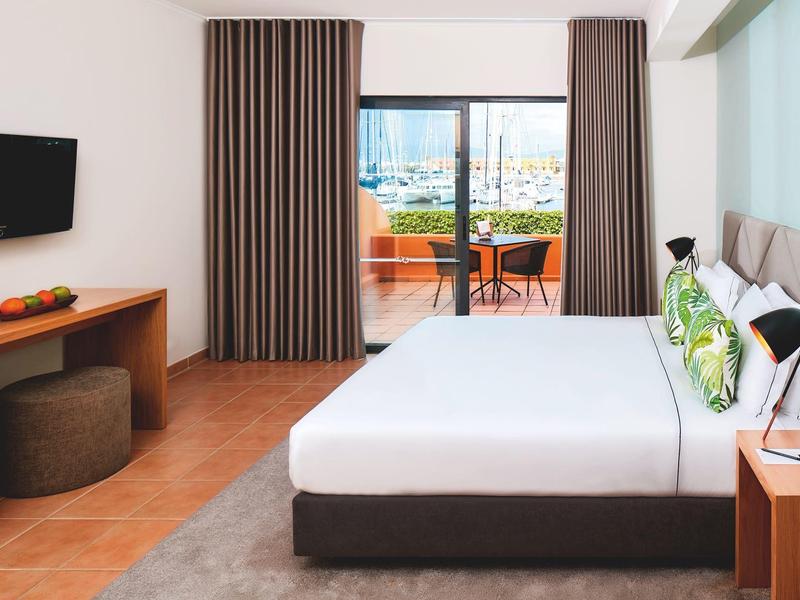 Hell eingerichtetes Schlafzimmer mit großem Bett, TV, Balkon und braunen Vorhängen.