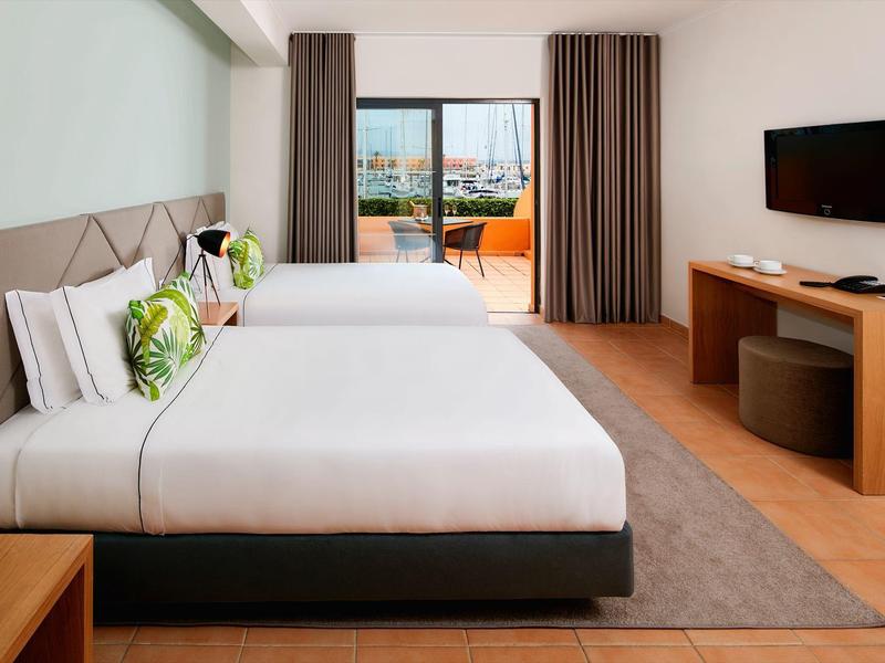 Modernes Hotelzimmer mit zwei Betten, großem Fenster, Flachbildfernseher und elegantem Holzdesign.