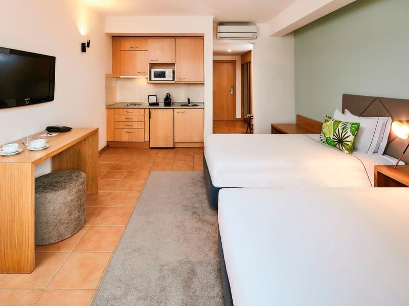 Hotelzimmer mit zwei Betten, Küchenzeile, Wand-TV und modernem, hellem Interieur.