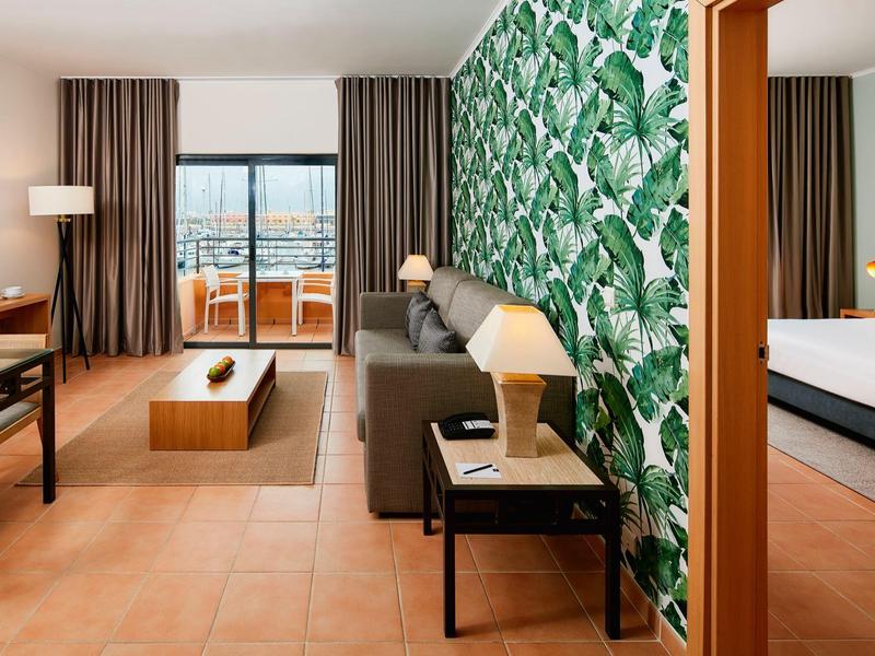 Modernes Hotelzimmer mit tropischer Wand, dunklem Sofa, Holzmöbeln und Balkon mit Meerblick.