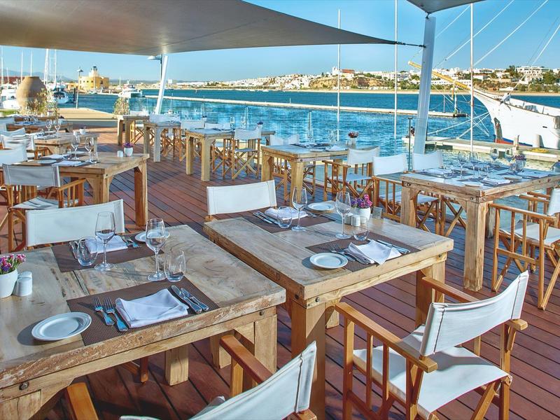 Freiluftrestaurant mit Holztischen, weißen Stühlen und Ausblick auf einen Hafen mit Booten.