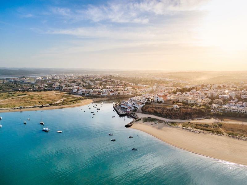 Küstenstadt mit Sandstrand, blauen Booten im Wasser und Sonnenlicht am Horizont.