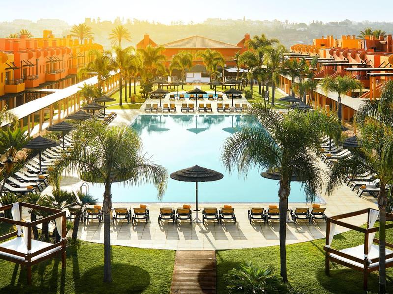 Großer Pool mit Liegestühlen, Sonnenschirmen und Palmen in einem sonnigen Resort.