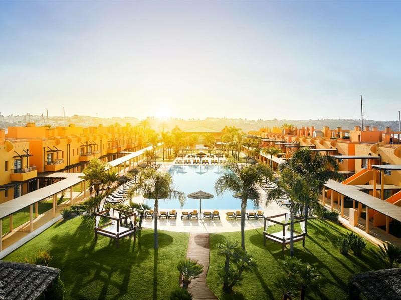 Sonnenuntergang über einem Resort mit Pool, Palmen und gelben Gebäuden auf beiden Seiten.