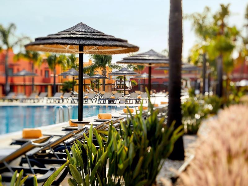 Poolbereich mit Liegestühlen, Sonnenschirmen und grünen Pflanzen an einem sonnigen Tag.