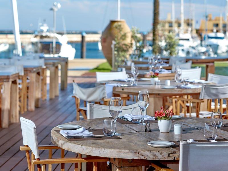 Freiluftrestaurant mit rundem Holztisch, weißen Stühlen und Blick auf den Hafen mit Booten.