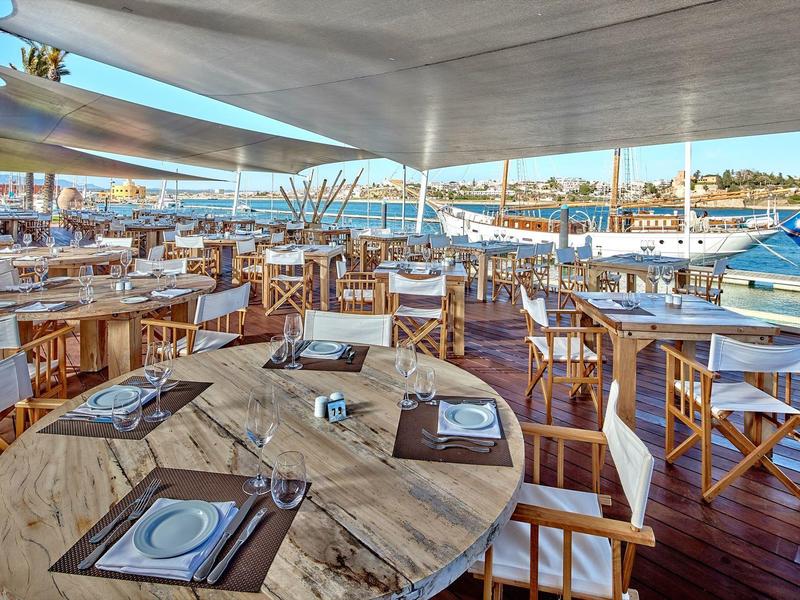 Offenes Restaurant mit Holztischen, weißen Stühlen und Blick auf Wasser und Stadt im Hintergrund.