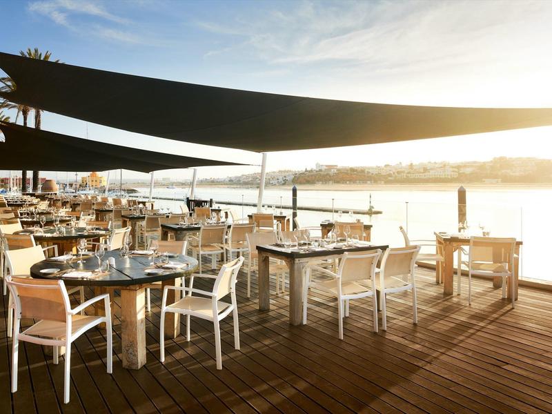 Sonnige Terrasse mit Holzboden, weißen Stühlen, Tischen und schwarzen Sonnensegeln am Wasser.