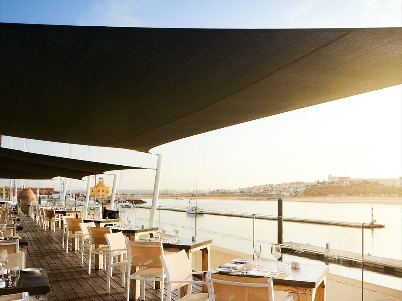 Terrassenrestaurant am Wasser mit weißen Stühlen, Holzboden und großer Sonnenmarkise.