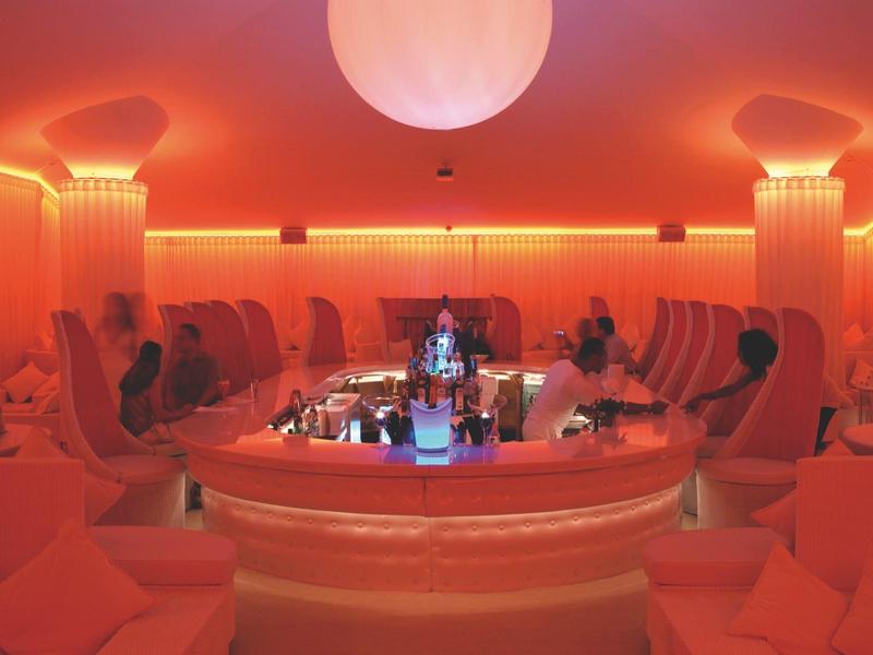 Innenraum einer Bar mit orangem Licht, runden Sitzecken und einer beleuchteten Theke in der Mitte.