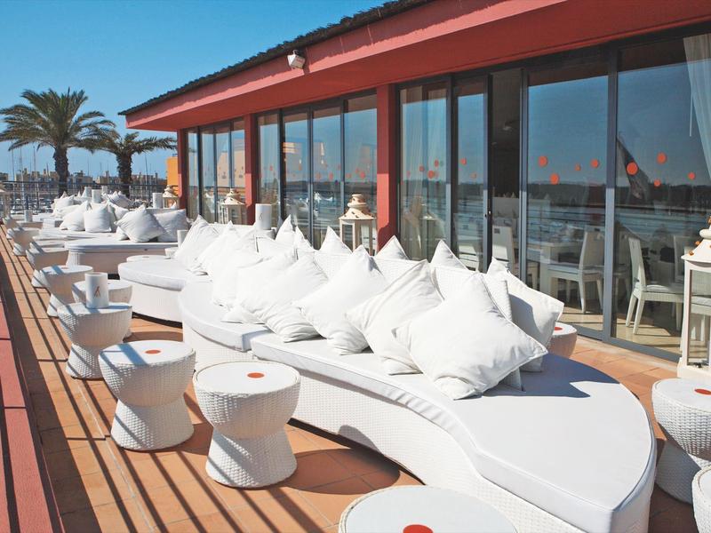 Moderne Dachterrasse mit weißen loungigen Sitzmöbeln, Holzboden und Glasfront bei Sonnenschein.