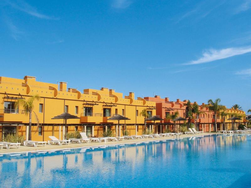 Großer blauer Pool vor gelben und orangenen Reihenhäusern unter blauem Himmel mit wenigen Wolken.