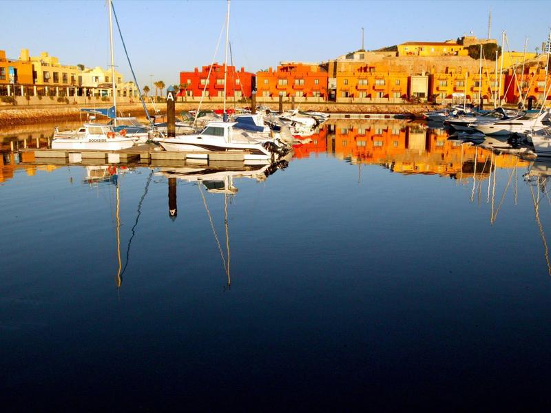 Hafen mit Segelbooten, ruhiges Wasser reflektiert bunte Häuser und Boote bei Sonnenlicht.