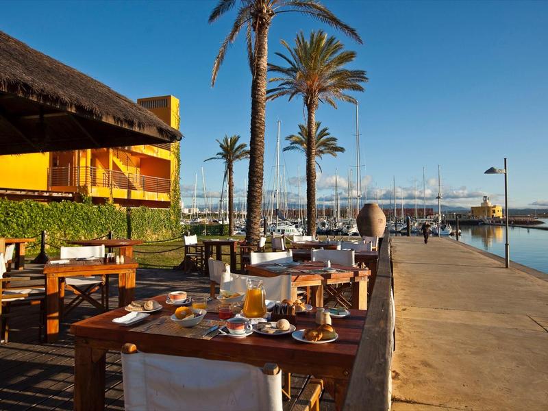 Terrassenrestaurant mit Holztischen, Palmblättern und Blick auf Hafen bei klar blauem Himmel.