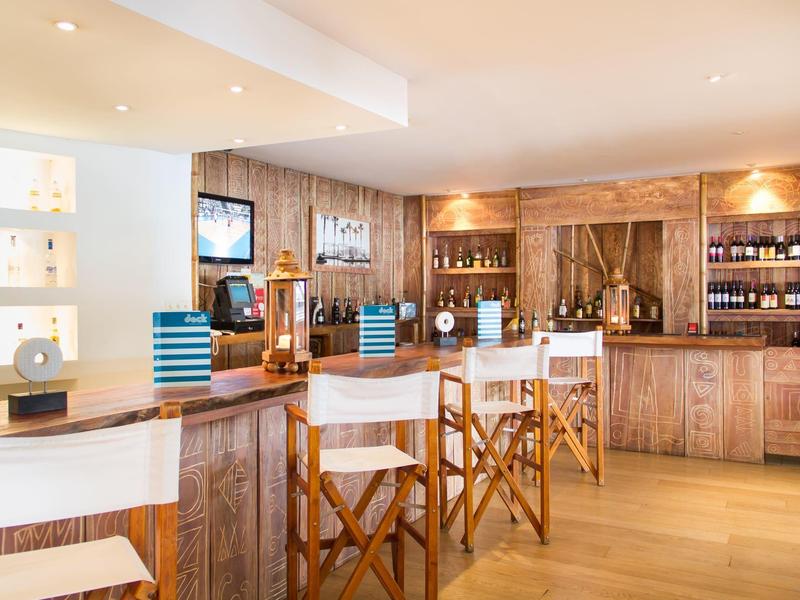 Helle Bar mit Holztheke, weißen Stühlen, Regalen voller Flaschen und warmem Licht.