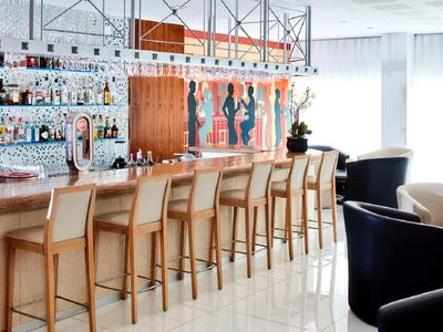 Moderner Barbereich mit Holztresen, sechs hellen Barhockern, Regalen voller Flaschen und heller, lichtdurchfluteter Atmosphäre.