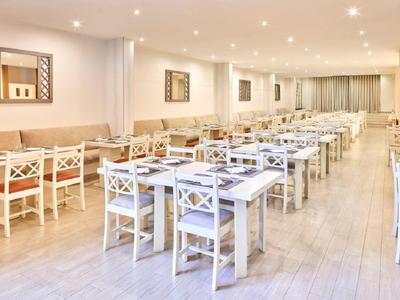 Heller, moderner Speisesaal mit weißen Holzstühlen und Tischen in mehreren Reihen.