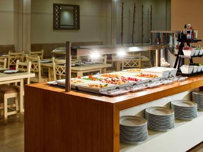 Buffet mit warmen Speisen auf Holztheke, Stapel Teller darunter, helle Holzmöbel im Hintergrund.