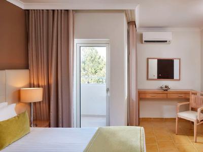 Helles Schlafzimmer mit weißer Bettwäsche, grünem Kissen, Vorhängen, Lampe und Holzschreibtisch.