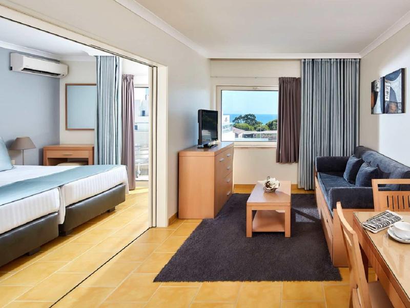 Hell eingerichtetes Hotelzimmer mit zwei Einzelbetten, Sofa, Fernseher und Esstisch, hellem Holzboden.