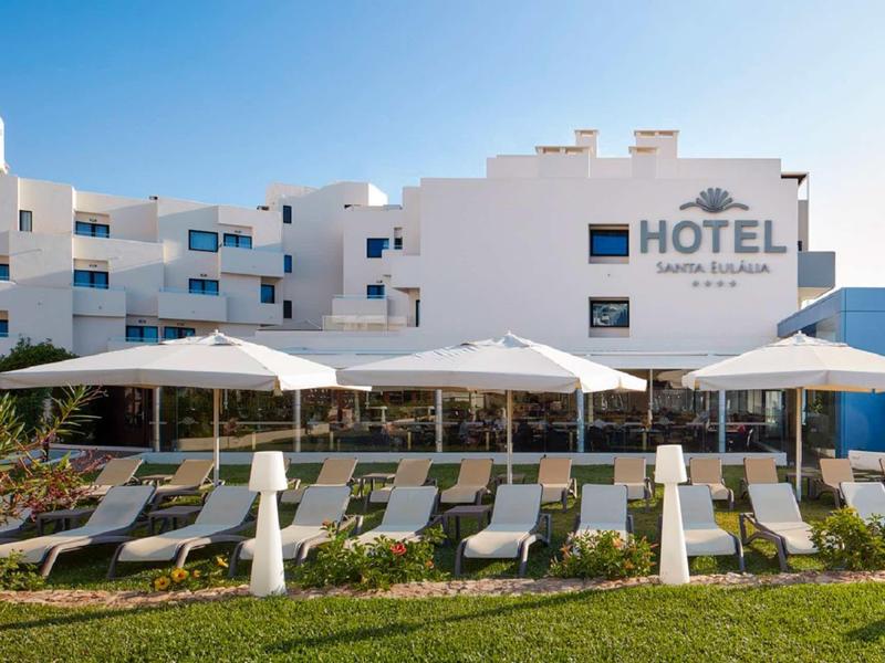 Hotelgebäude in Weiß mit Sonnenschirmen und Liegestühlen vor einer grünen Rasenfläche.