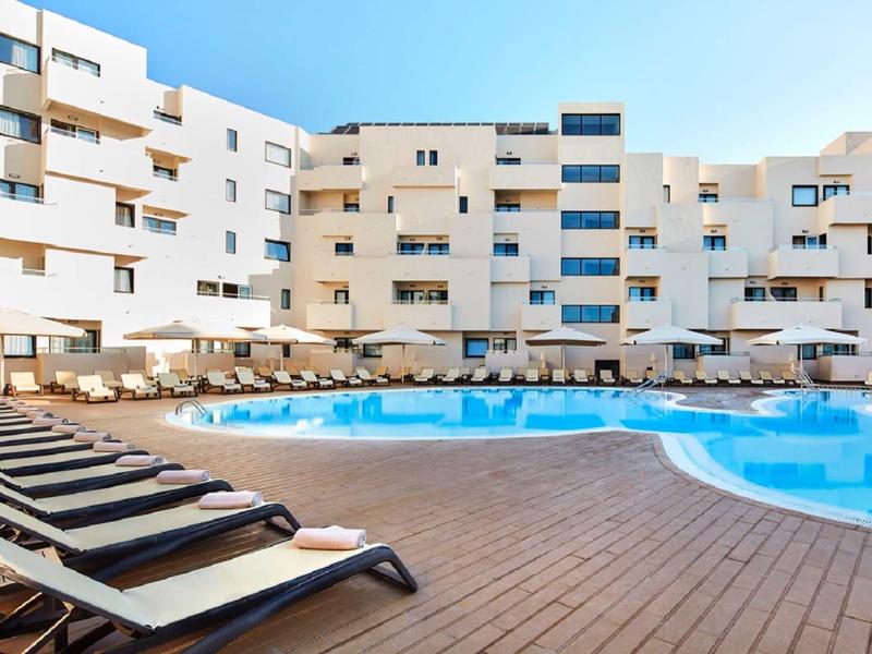 Moderner Hotelkomplex mit großem, geschwungenem Pool und Liegestühlen auf Holzterrasse bei Tageslicht.