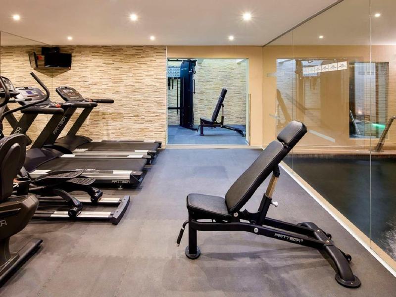 Fitnessraum mit Laufbändern, Trainingsbank und Spiegelwand, heller Steinwand und Schwimmbecken.