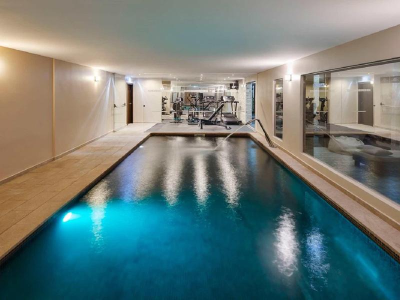 Innen-Pool mit blauem Wasser, beige Fliesenboden und großen Spiegeln an der rechten Wand.