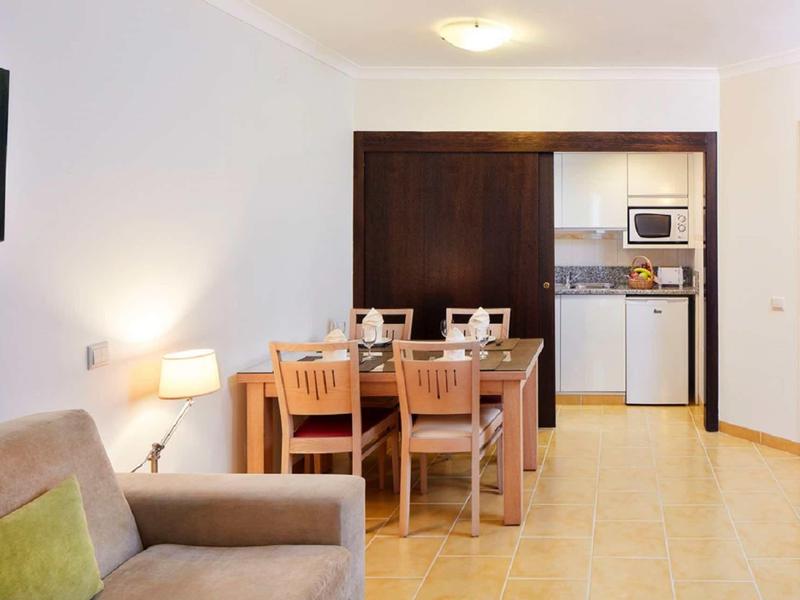 Wohnzimmer mit beige Couch, Holztisch mit vier Stühlen und kleiner Küchenzeile im Hintergrund.