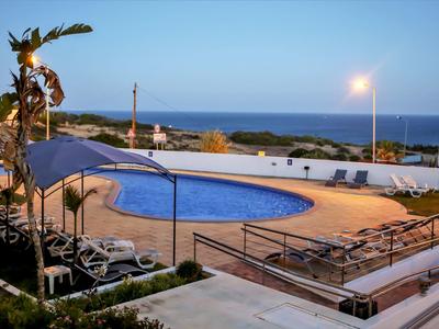 Schwimmbad mit blauer Wasserfläche, Liegestühlen, Palmen und Blick auf Meer bei Abenddämmerung.